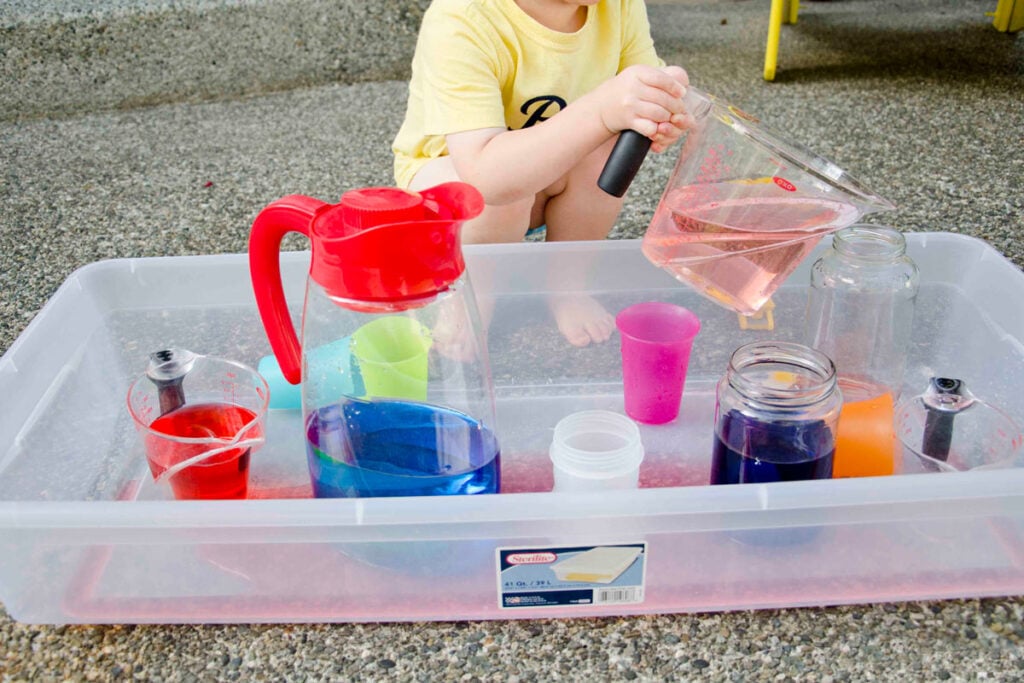 A child sits behind a storage container full of cups and water. He is lifting a large measuring cup full of light pink water.