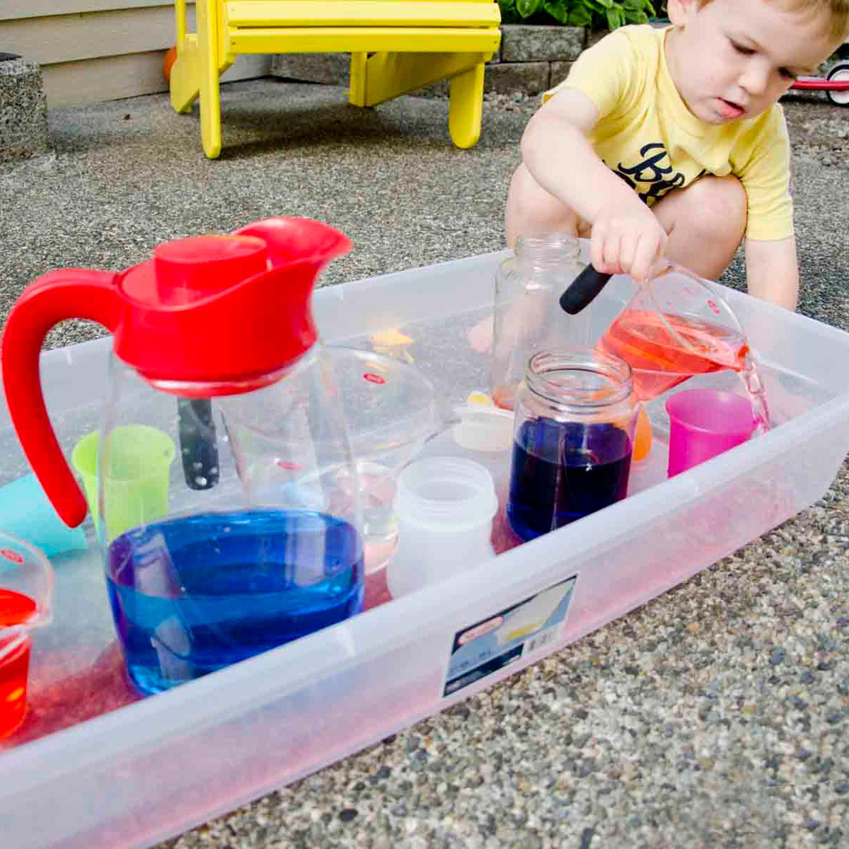 A child in a yellow shirt leans over a storage container full of cups and water. He's pouring orange water into a cup, and missing.