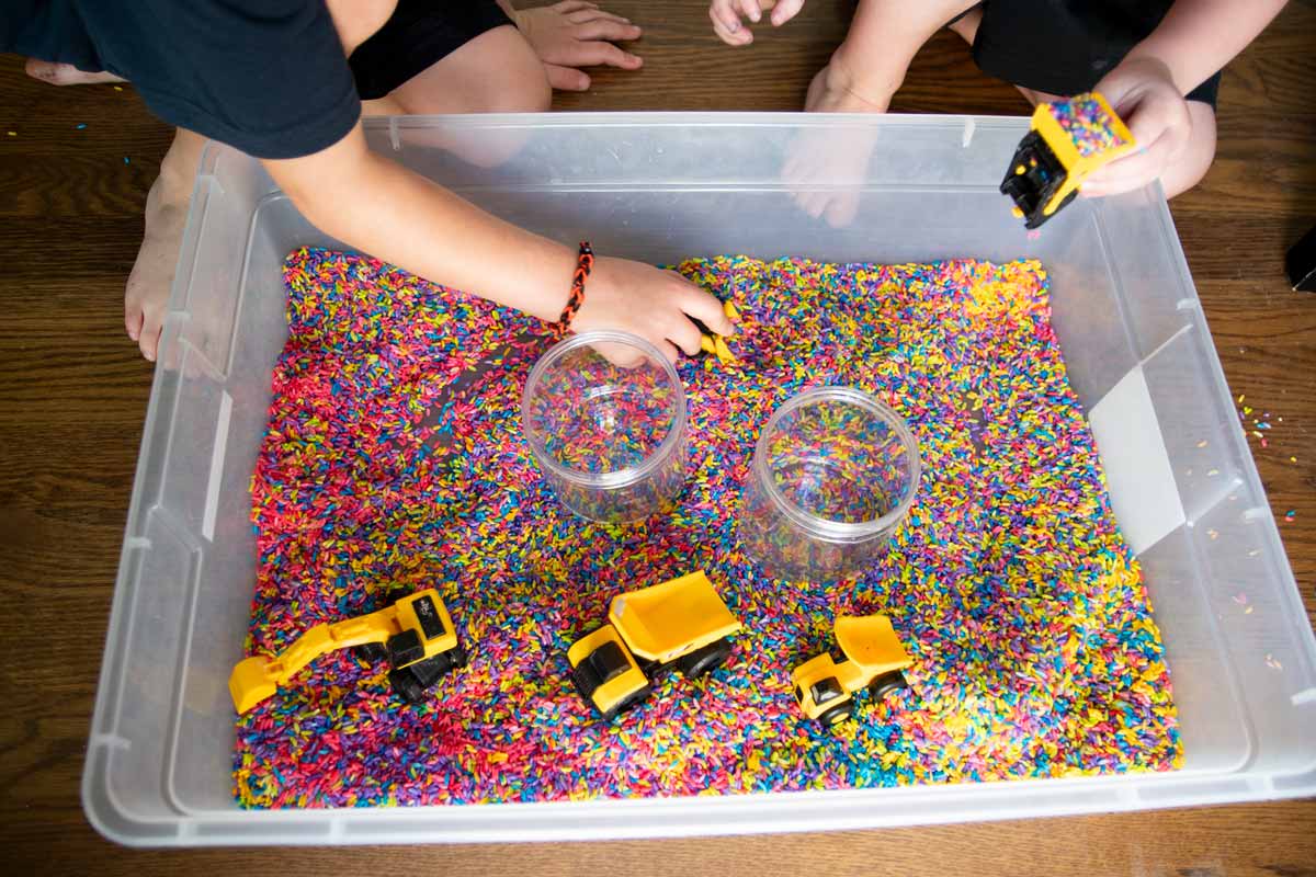 Two children sit on the floor behind a rainbow rice sensory bin. The children are playing with small construction trucks.