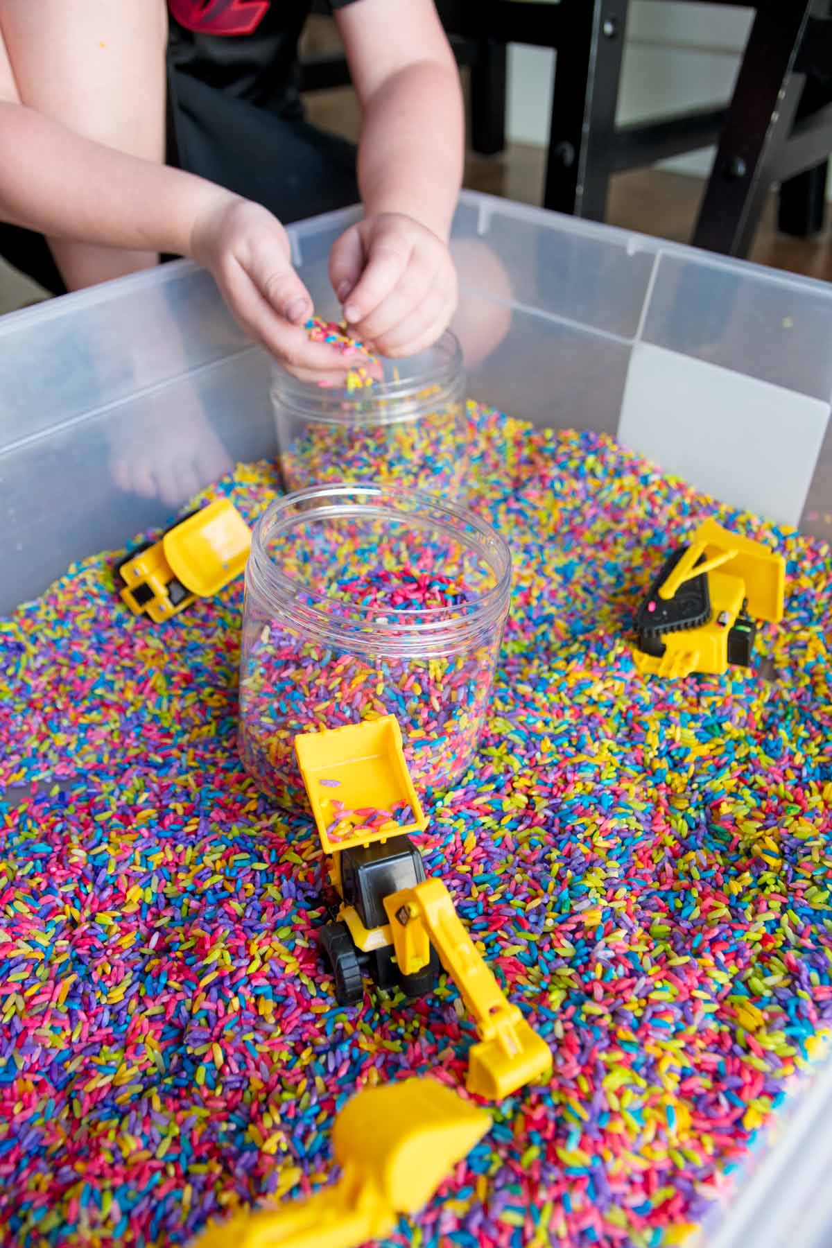 A child sits at the back corner of a rainbow rice sensory bin adding rice to jars. Construction trucks are present.