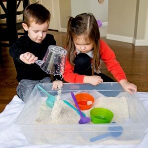 Two kids play with a rice sensory bin. One is dumping a measuring cup of rice back into the bin.