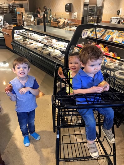 Three kids in a grocery store. One child is standing and two are in the cart. All three have donuts.