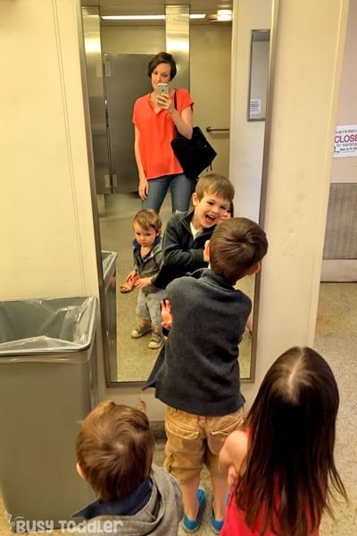 A parent takes a selfie in a mirror of 3 kids playing in a public bathroom.