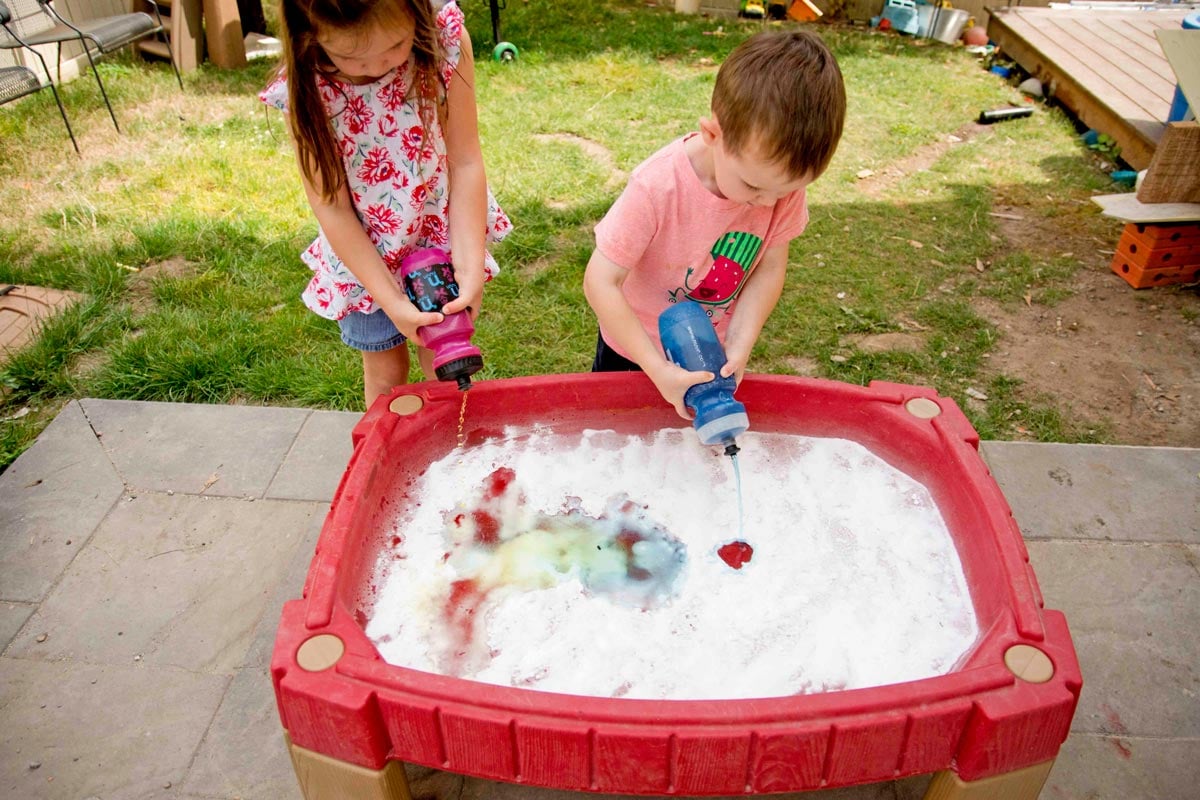 An overhead shot of two children outside at a water table full of baking soda using squirt bottles to squeeze in colorful vinegar.