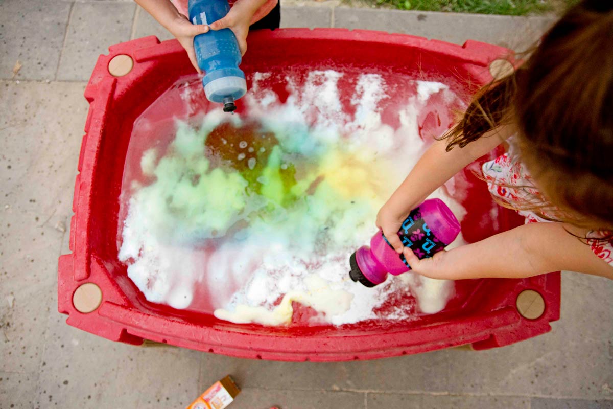 Overhead shot of a sand table full of baking soda and vinegar, fizzing. Two kids hold squirt bottles.