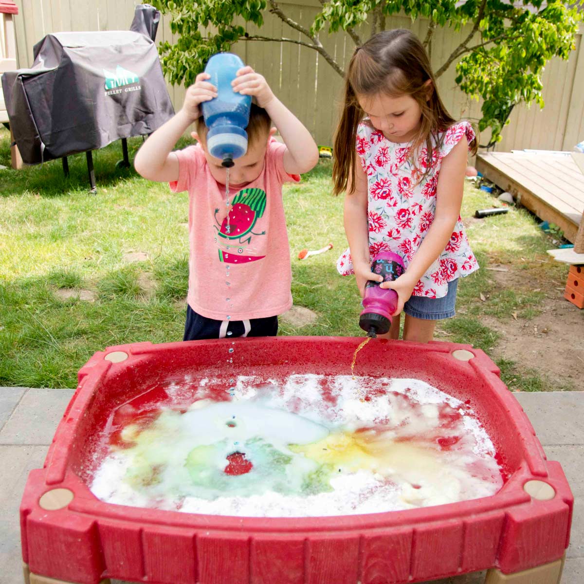 Two children stand in a backward at a water table full of baking soda using squirt bottles to squeeze in colorful vinegar.