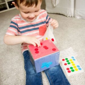 A child smiles while placing a red dot sticker onto a box covered in construction paper. He's sorting stickers onto each side of the box.