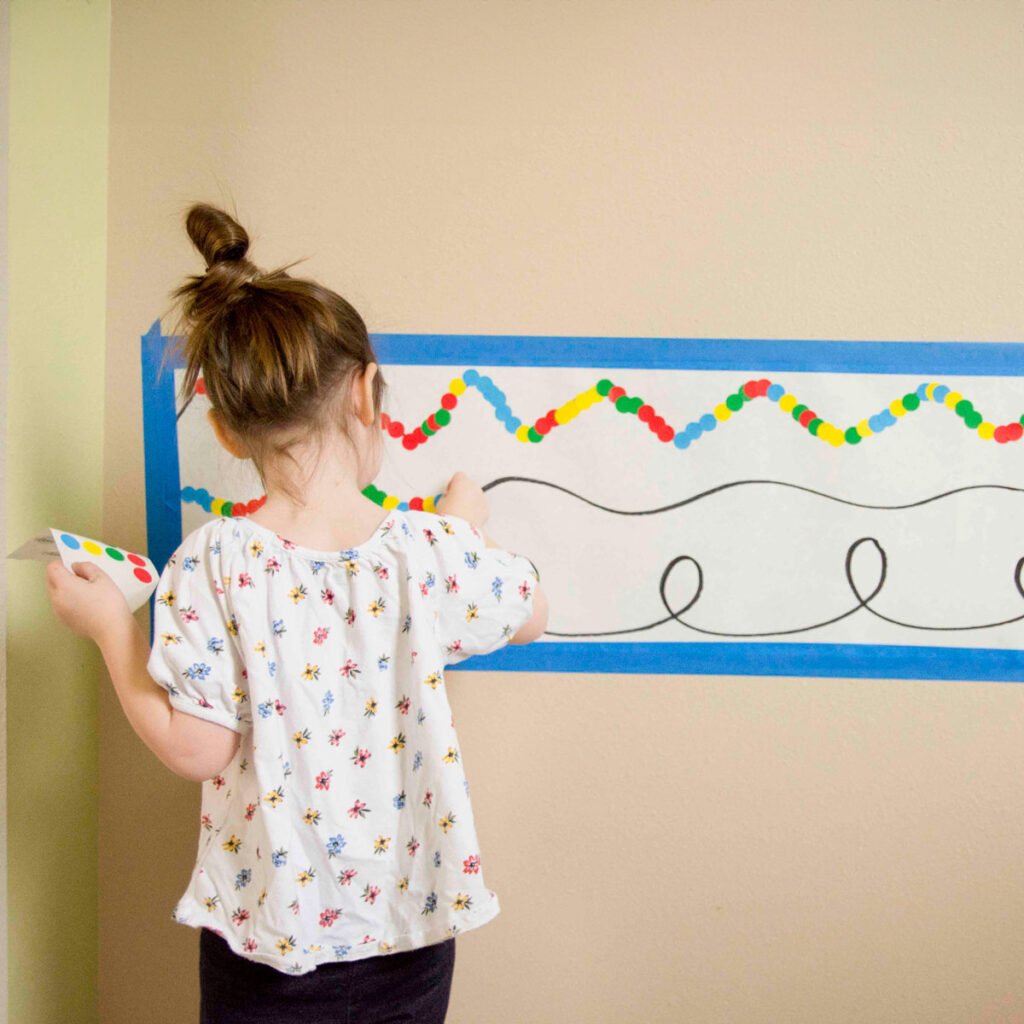 A child stands at a wall with a white piece of long paper on it. There are black lines. The child is placing stickers on the lines.