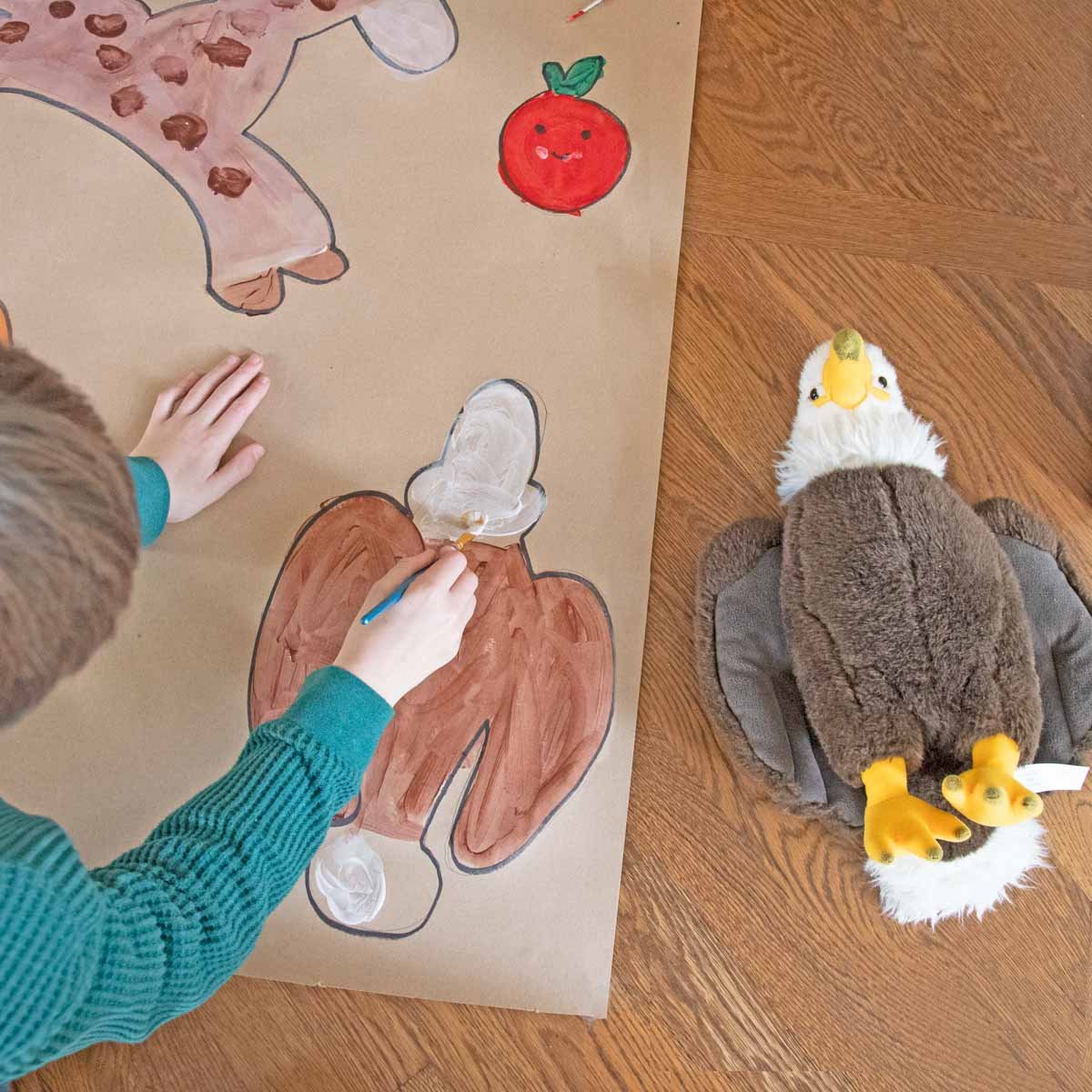 A child paints a portrait of their eagle stuffed animal. There is a smiling apple picture above him.