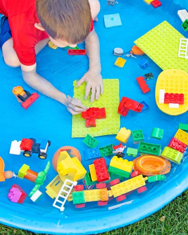 A child in a blue kiddie pool plays with LEGO Duplo bricks.