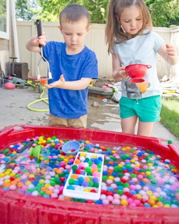 Two children play with a water table full of water and pom poms