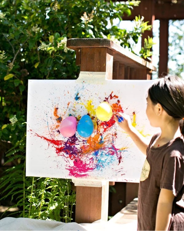 A child stands at a canvas popping balloons full of paint.