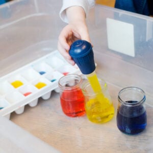 A child's hand squeezes a turkey baster, filling it with yellow water. Jars of red, yellow, and blue water are present, along with an ice cube tray.