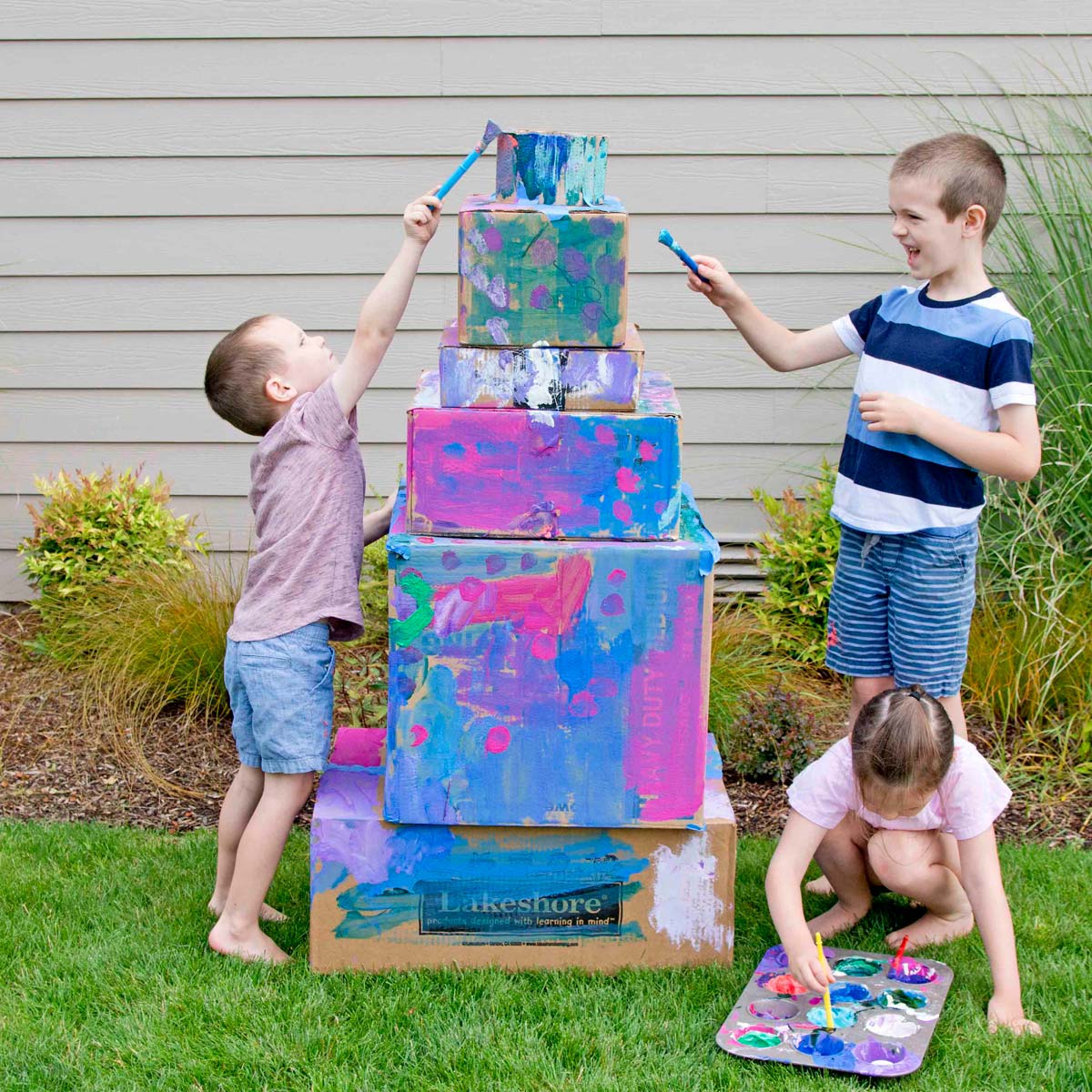 Three kids are outside painting a cardboard box wedding cake. One child is laughing, one is reaching up, and one is putting more paint on their brush.