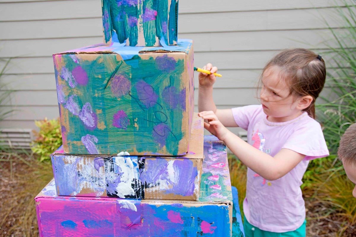 A child is painting a stack of boxes with turquoise, purple, and pink paint.