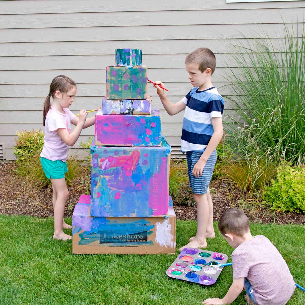 Three kids are painting a box cake outside. The cake has six tiers. They are using paint from a muffin tin which is sitting on grass.
