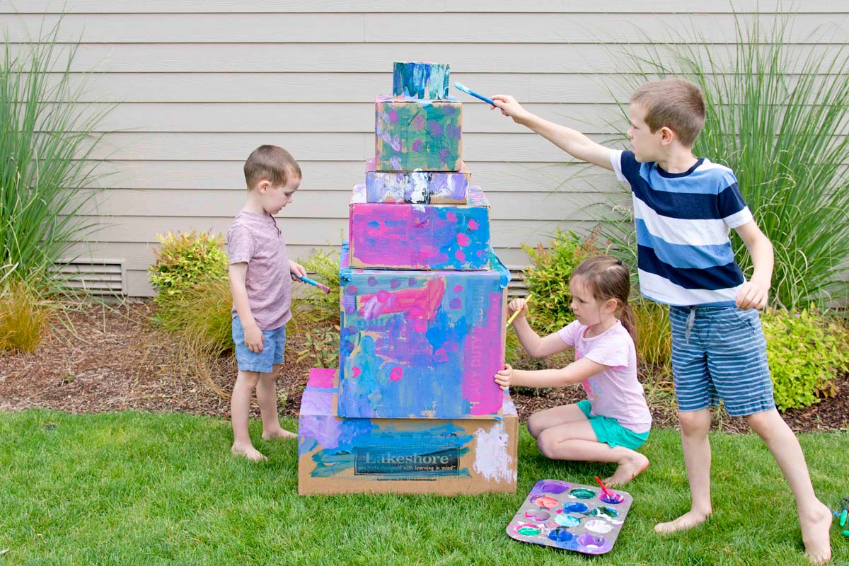 Three kids are outside painting a six tier box wedding cake. The kids are 3, 5, and 7 years old.