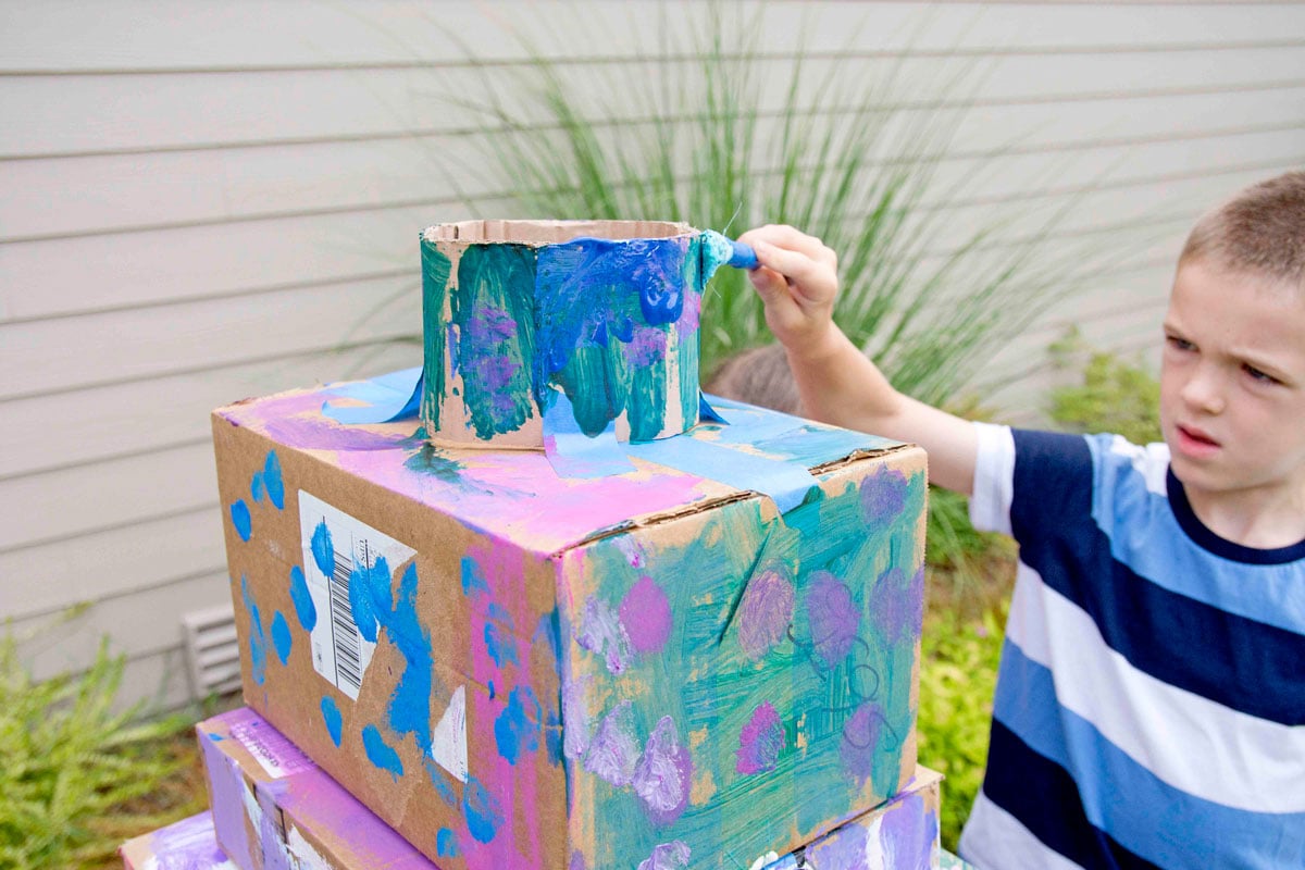 A child paints the top piece of a box wedding cake. He is using turquoise paint on cardboard.